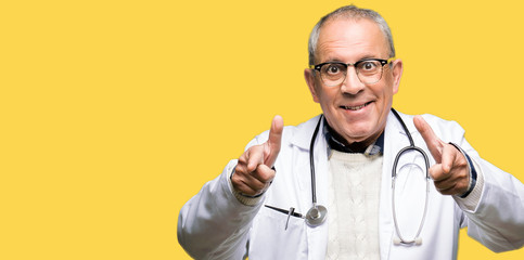 Handsome senior doctor man wearing medical coat pointing fingers to camera with happy and funny...