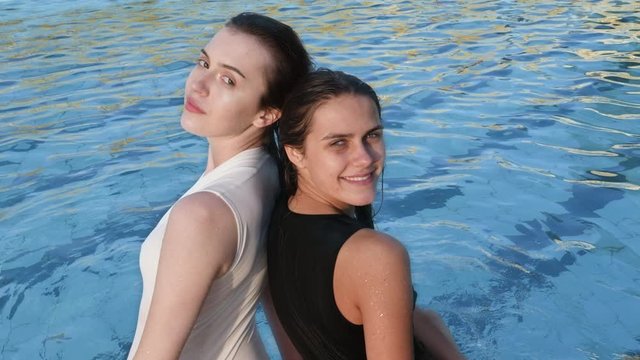 Two young women are sitting on the edge of the pool with clear blue water. Girls in a bikini.