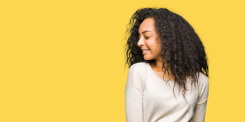 Young beautiful girl with curly hair wearing casual sweater looking away to side with smile on face, natural expression. Laughing confident.