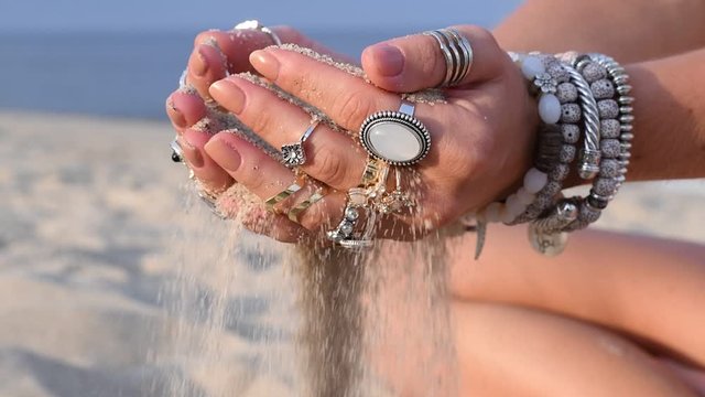 Beautiful woman's hands with accessories is pouring sand running through fingers at the beach in slow motion