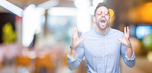 Young handsome man wearing sunglasses over isolated background crazy and mad shouting and yelling with aggressive expression and arms raised. Frustration concept.