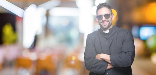 Young Christian priest wearing sunglasses over isolated background happy face smiling with crossed arms looking at the camera. Positive person.