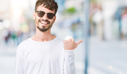 Young handsome man wearing sunglasses over isolated background smiling with happy face looking and pointing to the side with thumb up.