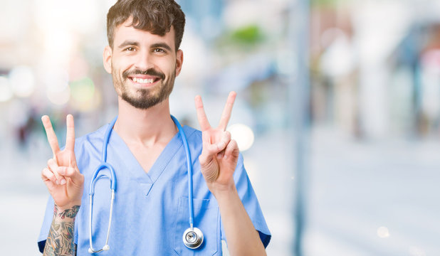 Young handsome nurse man wearing surgeon uniform over isolated background smiling looking to the camera showing fingers doing victory sign. Number two.