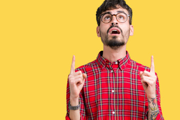 Young handsome man wearing glasses over isolated background amazed and surprised looking up and pointing with fingers and raised arms.