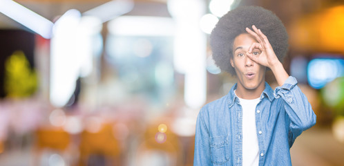 Young african american man with afro hair doing ok gesture shocked with surprised face, eye looking through fingers. Unbelieving expression.