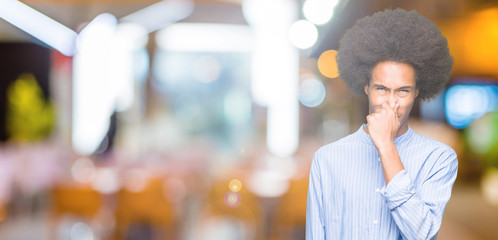 Young african american man with afro hair smelling something stinky and disgusting, intolerable smell, holding breath with fingers on nose. Bad smells concept.