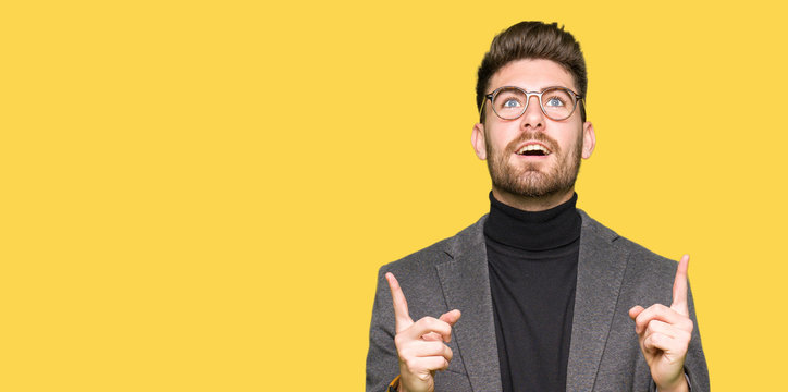 Young handsome business man wearing glasses amazed and surprised looking up and pointing with fingers and raised arms.