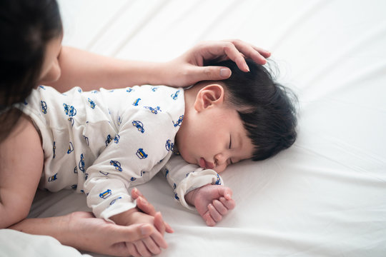 Top View Of Asian Mother Is Touching Her Cute Sleeping Newborn Baby Head On Bed. She Smiling And Putting Her Hand A Warm Touching On Kid Hair With Love In White Bedroom At Home. Happy Family Concept.