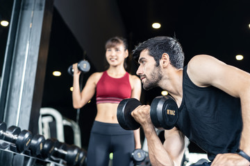 Fototapeta na wymiar Young Caucasian handsome and pretty Asian girl are lifting the weights in gym or fitness club. They both have very strong and good body shape. Seen side of the man is looking straight in the mirror.