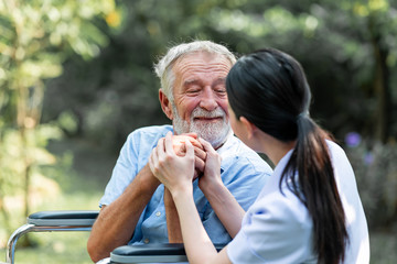 Naklejka premium Caring nurse with senior man sitting on wheelchair in gaden. Asian woman, caucasian man. Asking questions.