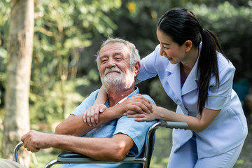Fototapeta premium Caring nurse with senior man sitting on wheelchair in gaden. Asian woman, caucasian man. Morning talk.