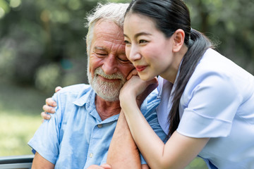Caring nurse with senior man sitting on wheelchair in gaden. Asian woman, caucasian man. Close up, comforting smile.