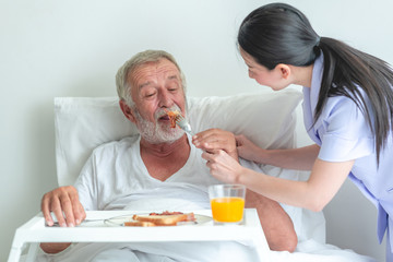 Senior man in bed having breakfast with nurse. Caucasian man with asian woman.  Feeding him.