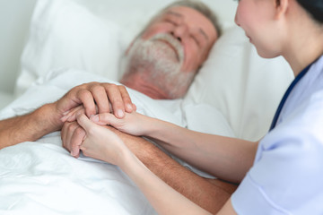 Fototapeta na wymiar Senior man in bed with nurse in retirement home. Caucasian male with asian woman. Holding hands while he sleeps.