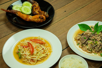 Most popular Thai northeast food with papaya salad, grilled chicken wings, minced pork and liver salad with sticky rice on wooden table background.