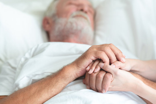 Senior man in bed with nurse in retirement home. Caucasian male with asian woman. Holding hands, close up shot.