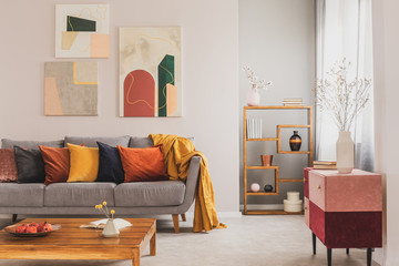 Yellow, orange, black and brown pillows on comfortable grey scandinavian sofa in bright living room interior with abstract paintings on the wall - obrazy, fototapety, plakaty
