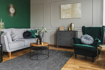 Emerald green wing back chair with pillow in grey living room interior with wooden commode © Photographee.eu