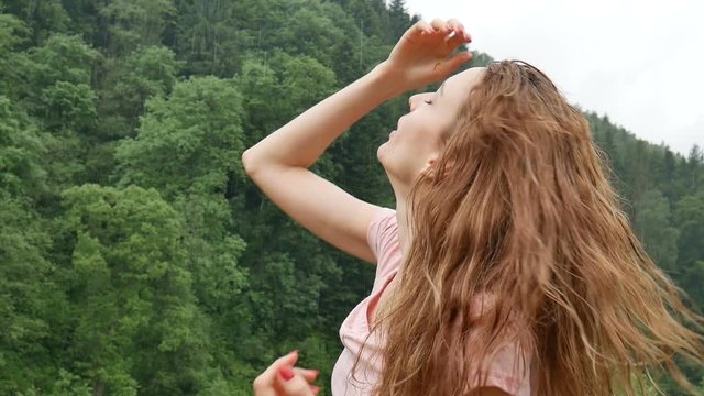 Close up of sexy young woman with blonde wet hair and pink dress breathing during cold summer rain outdoors in the mountains on green forest background