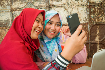 Fototapeta premium Happy and pretty Asian Indonesian girls in traditional Islamic hijab head scarf taking selfie together having fun at cafe in friendship and social media concept