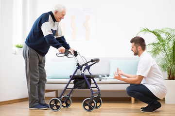 Elderly grandfather with walker trying to walk again and helpful male nurse supporting him