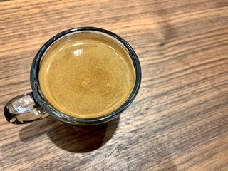  A cup of hot black coffee with bubbles in a glass cup with the light and shadow on wooden table background.