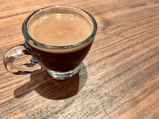 A cup of hot black coffee with bubbles in a glass cup with the light and shadow on wooden table background.