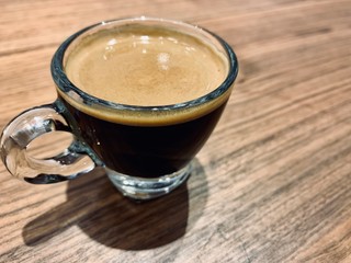  A cup of hot black coffee with bubbles in a glass cup with the light and shadow on wooden table background.