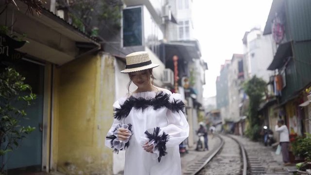 Beautiful and young fashion mestizo girl walking along the asian street with railway between old buildings. Journey concept in Vietnamese tourist destination. Cute female in white fashionable dress.