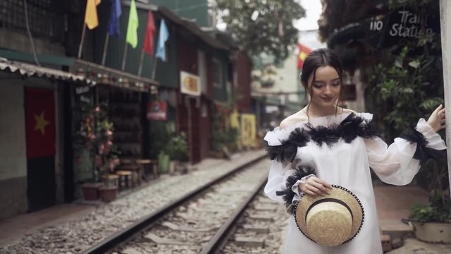 Beautiful and young fashion mestizo girl walking along the asian street with railway between old buildings and holding straw hat in hands. Journey concept in Vietnamese tourist destination in Hanoi. 