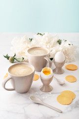 Fototapeta na wymiar Soft-boiled eggs with liquide orange yolk in ceramic egg cups, two cups of coffee with ceramic spoons and thin crispy corn chips on background of beautiful white peony. Breakfast concept. Copy space.