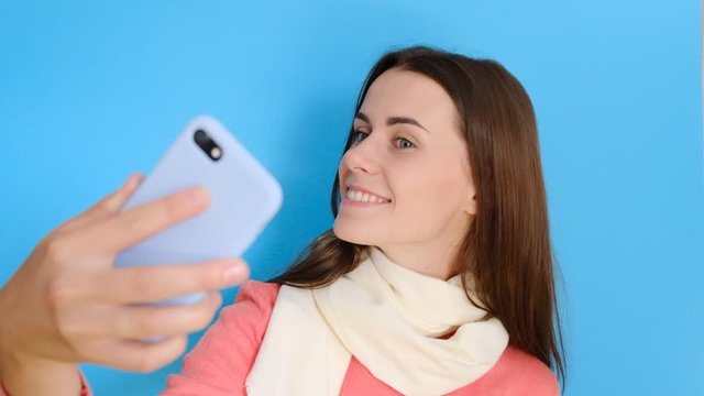 Happy pretty girl smiles, takes selfie portrait on cell phone, enjoyed free time, makes pics for social networks, stylish scarf and rosy jumper, smiles at camera isolated over blue background