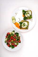 Sandwiches with poached eggs and salad with avocado, cucumbers, cherry tomatoes, spinach leaves, parsley and seeds. Breakfast food on a white background. Top view.