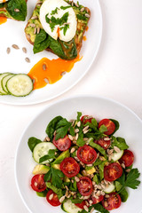 Sandwiches with poached eggs and salad with avocado, cucumbers, cherry tomatoes, spinach leaves, parsley and seeds. Breakfast food on a white background. Top view.