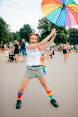 Obraz premium bisexual girl with lgbt gay pride flag on her face, colorful umbrella, bag and mismatched socks posing on street with people on background.