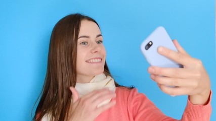 Portrait of beautiful girl smiles, takes selfie portrait on cell phone, enjoyed free time, makes pics for social networks, stylish scarf and rosy jumper, smiles at camera, isolated over blue backgroun