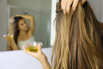 Young woman applying olive oil mask on hair in front of a mirror. Hair care concept. Focus on hair.