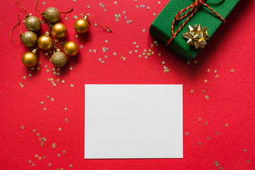 Christmas composition. White blank paper, wrapped gift, toys on red background with golden confetti. new year concept. Greeting card, xmas celebration 2020. Flat lay, top view, copy space, mockup