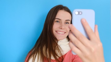 Positive young girl smiles, takes selfie portrait on cell phone, enjoyed free time, makes pics for social networks, stylish scarf and rosy jumper, smiles at camera, isolated over blue background