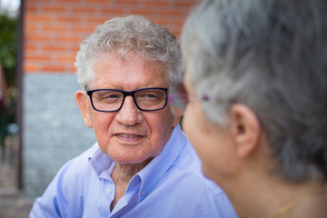 Portrait of an older man with grey hair and eyeglasses, smiling at his wife in outdoor scenery. The woman is out of focus.
