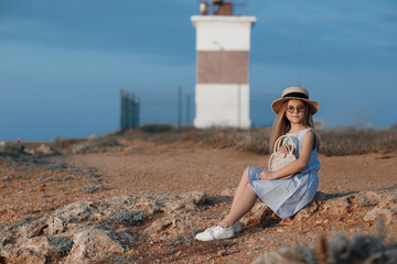 A girl of 10-11 years spends time alone on a rocky shore near the blue ocean on open air in the summer. Cute girl in a straw hat on a rocky coast. Little girl in a blue dress on a background of blue s