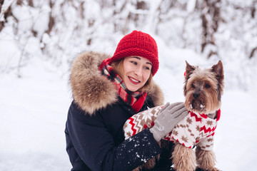 Woman spending time with a wonderful pet outdoors. Girl in a red hat and scarf playing with snow in the forest with a dog. snowy winter. Yorkshire Terrier in a Christmas costume plays in the snow.