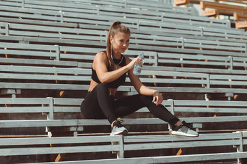 Tired woman in headphones resting and drinking water on grandstand