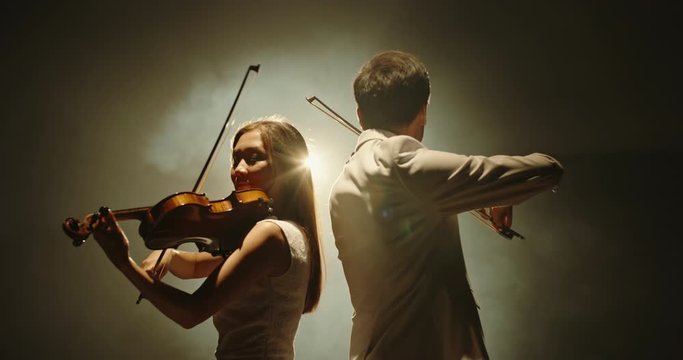 Male and female violin players having an amazing solo, standing spine-to-spine on stage, spotted by light on smoked black background 4k footage