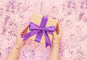 Child's hands holding giftbox tied with purple color ribbon on pink background with glitter. Flat lay style.