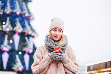 cute young girl wearing hat and pink sweater. christmas market lights bokeh background. Woman drinking hot chocolate in winter morning in christmas market.