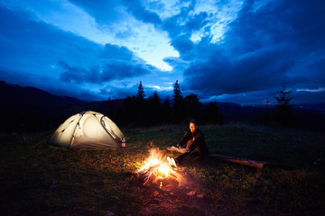 Fototapeta na wymiar Young woman traveller enjoying at night camping in the mountains, sitting near burning campfire and illuminated tourist tent under beautiful evening cloudy sky. Tourism, outdoor activity concept