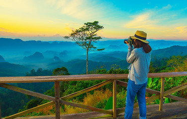 Fototapeta na wymiar Beautiful panoramic view with young asia woman taking landscape pictures with DSLR camera feeling happy and wearing hat stand resting on misty mountain, Doi Ta Pang view point, Chumporn, Thailand.