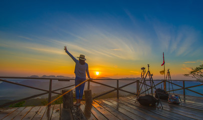 Fototapeta premium Beautiful panoramic view with young asia woman raise their hand up victory sign show feeling very happy wearing hat stand resting on misty mountain, Doi Ta Pang view point, Chumporn, Thailand.